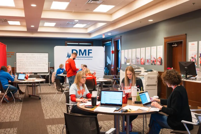 Volunteers work on laptops at round tables inside a DMF event space with a whiteboard and banner visible