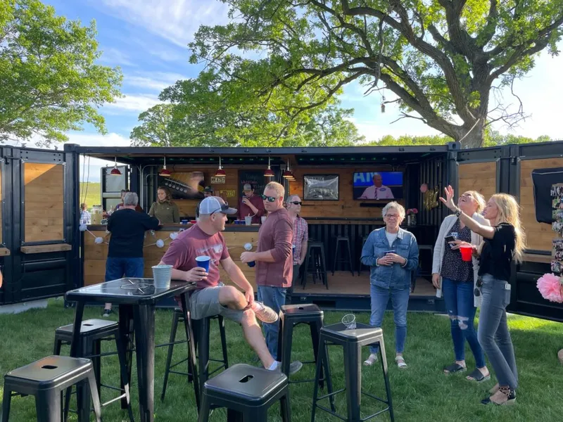 Guests socializing outdoors around a bar built inside a shipping container on a grassy lawn
