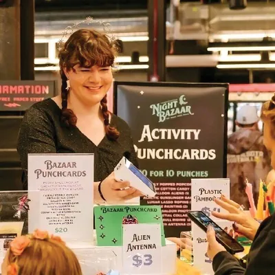 Smiling vendor at a Night Bazaar activity punchcard booth hands cards to a customer