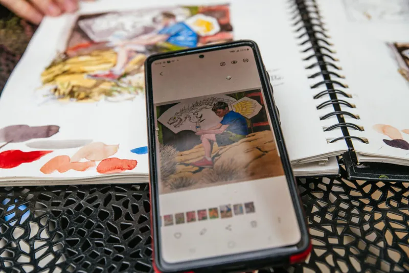 Smartphone resting on open sketchbooks with watercolor swatches, displaying a photo of an artist painting
