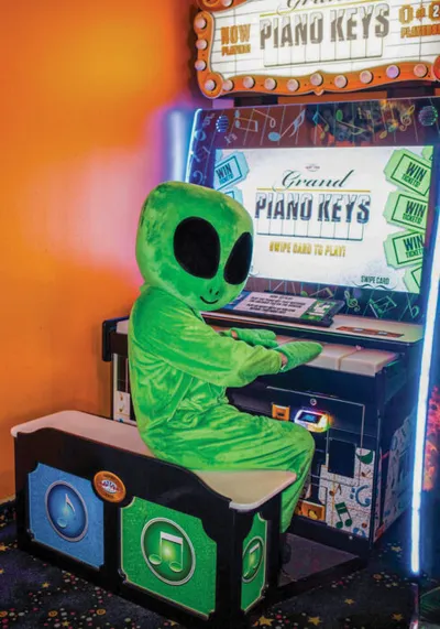 Person in green alien costume sitting at Grand Piano Keys arcade machine