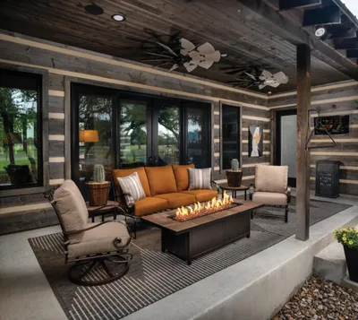 Covered patio with orange sofa, swivel chairs, and rectangular gas fire pit table beside a rustic log exterior