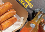 Golden fried spring rolls in a takeout box beside a small bottle of hot sauce