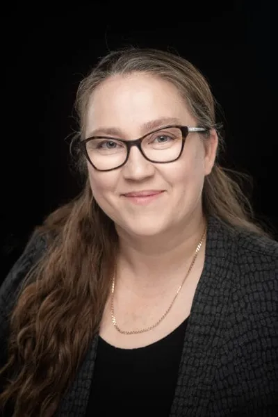 Woman with long brown hair, glasses, and a gold chain necklace smiling against a dark background