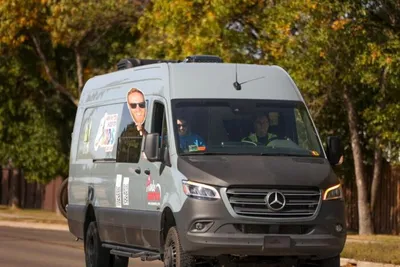 Gray Mercedes-Benz cargo van with a man's portrait decal driving on a tree-lined street