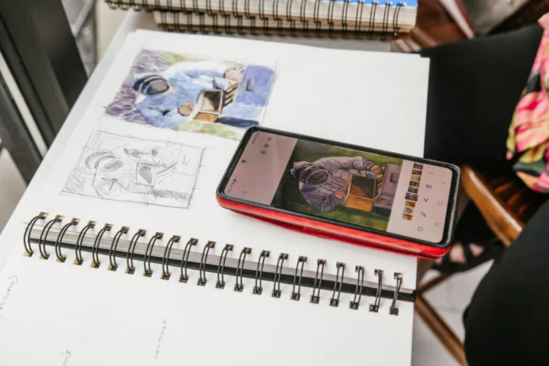 Open spiral sketchbook with pencil sketches and watercolors beside a smartphone displaying a painting