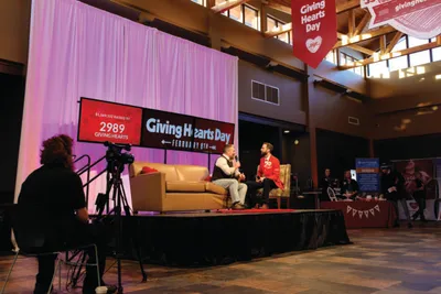 Two people on a stage during a Giving Hearts Day event, with a camera operator filming in the foreground