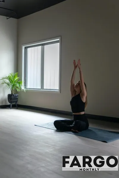 Woman in black activewear seated on a yoga mat, arms raised overhead in a bright studio