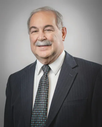 Older man with gray hair and mustache smiling in a dark pinstripe suit and patterned tie