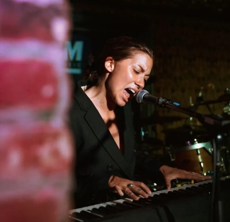 Woman in black blazer singing into a microphone while playing keyboard on a dimly lit stage