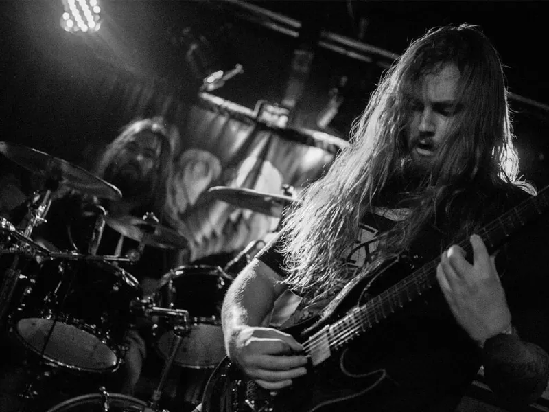Long-haired guitarist plays electric guitar on a darkened stage, drummer visible behind