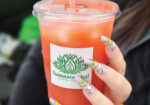 Hand holding a large iced pink-orange drink in a clear plastic cup with a green logo
