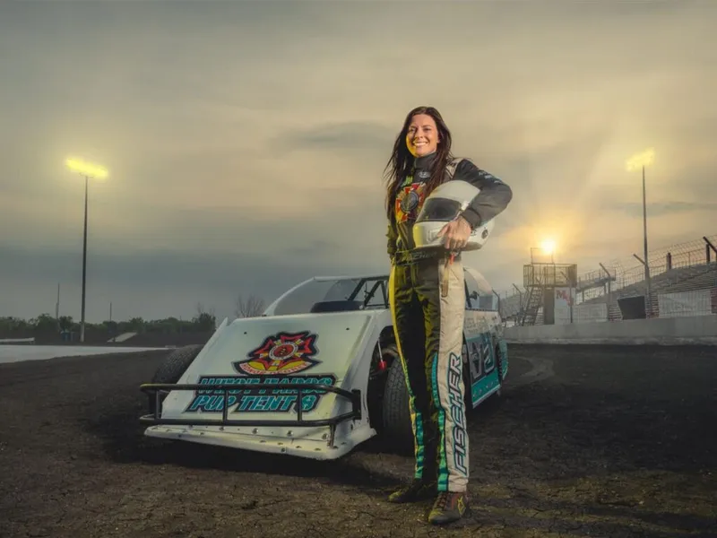 Smiling woman in a racing suit holds a helmet beside a dirt track race car at dusk