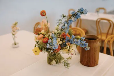 Glass jar with blue, orange, and white wildflowers beside a brown wooden vase on a white table