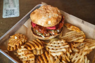 Sauced fried chicken sandwich on a brioche bun with waffle fries on a metal tray