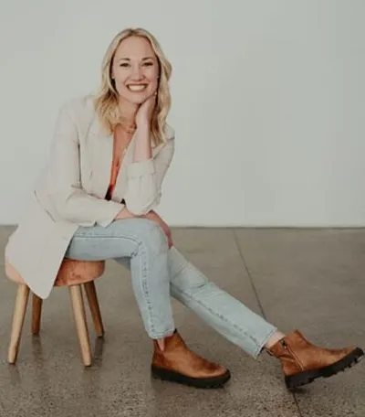 Smiling blonde woman seated on a stool, wearing a cream blazer, jeans, and brown boots