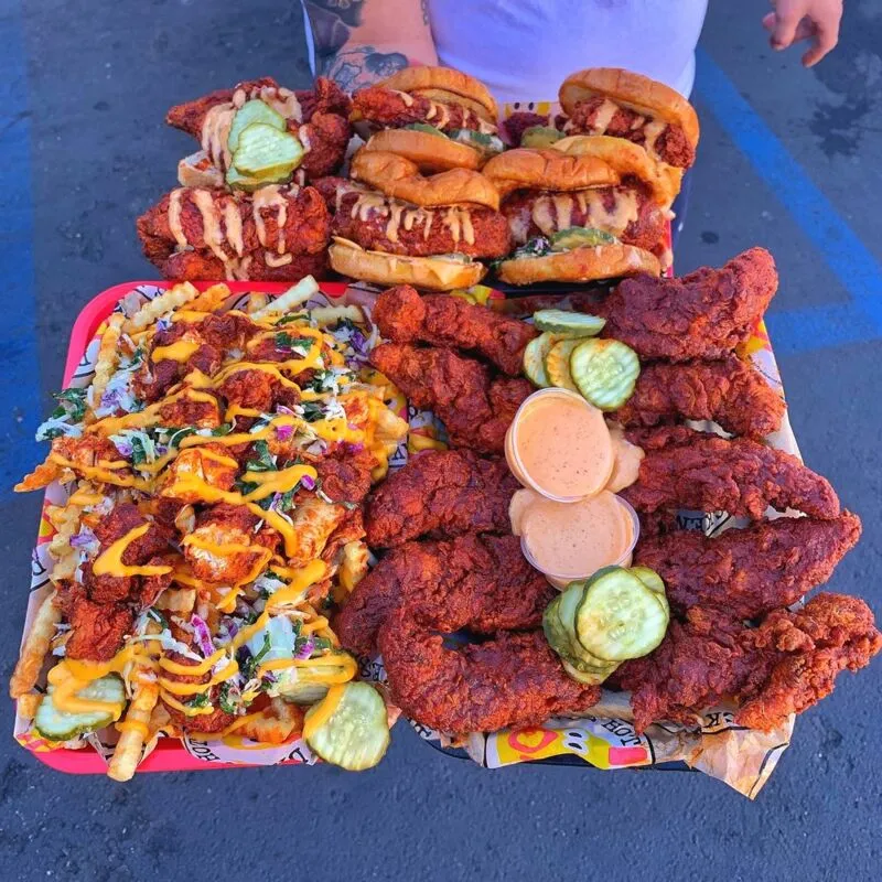 Large spread of spicy fried chicken tenders, sandwiches, loaded fries, pickles, and dipping sauces on red trays