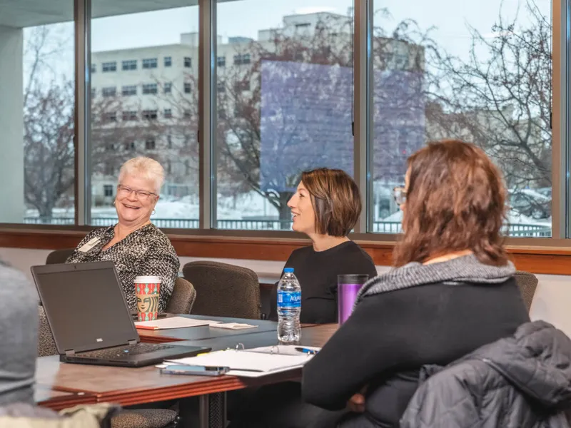 Three women smiling and conversing around a conference table with a laptop and beverages