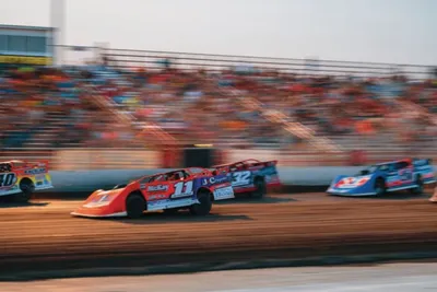 Dirt track race cars numbered 11 and 32 speeding past packed grandstands