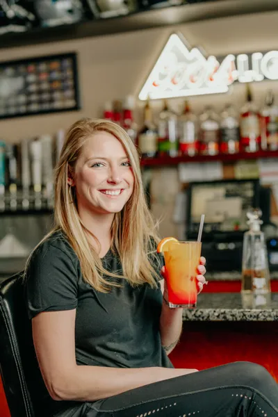 Smiling woman holding a layered red-orange cocktail with a straw at a bar