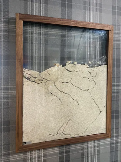 Framed sand art piece with wavy patterns hung on plaid wallpaper