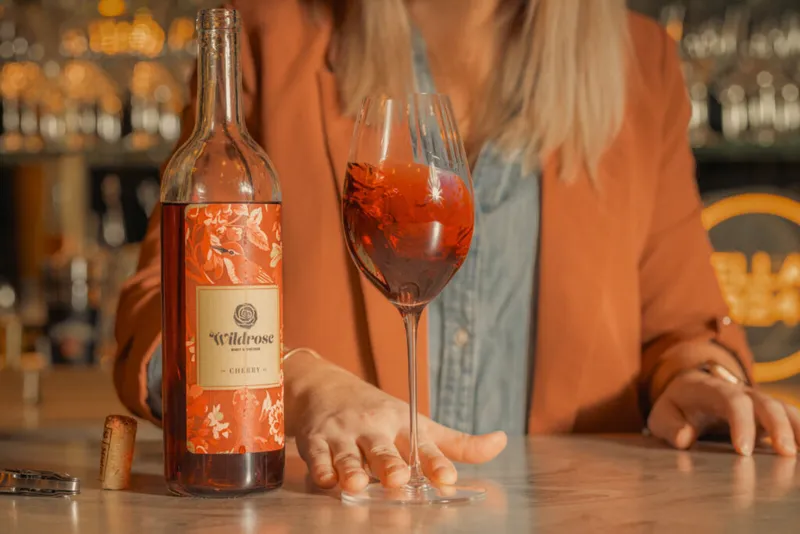 Wildrose Cherry wine bottle beside a filled glass on a bar, with a cork and woman's hand nearby