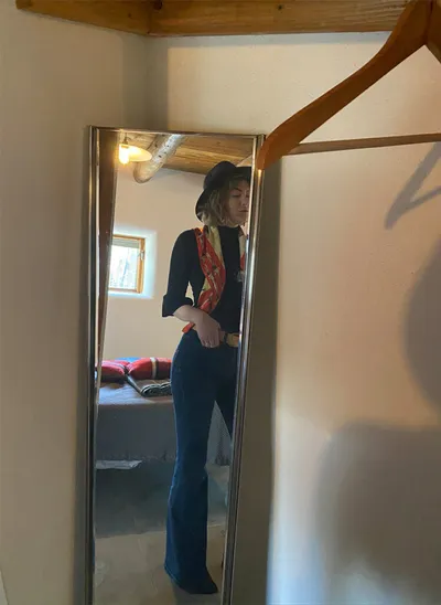 Woman in black top, flared jeans, and hat poses in a full-length mirror selfie