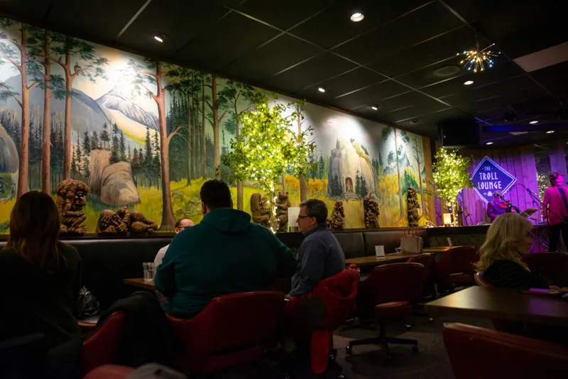 Guests seated facing a forest mural with troll sculptures at the Troll Lounge, live band performing on a purple-lit stage