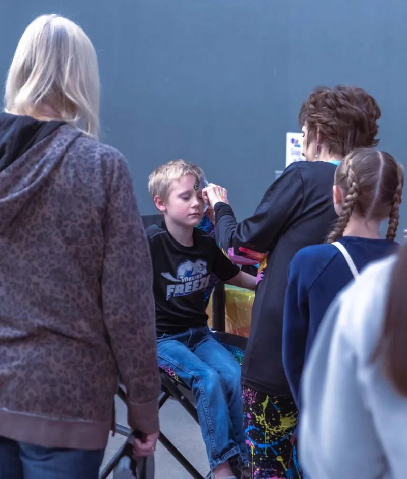 Face painter applying design to a boy seated on a folding chair at an indoor event
