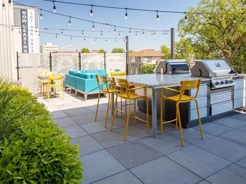 Rooftop patio with yellow bar stools, granite counter, gas grills, teal sofa seating, and string lights