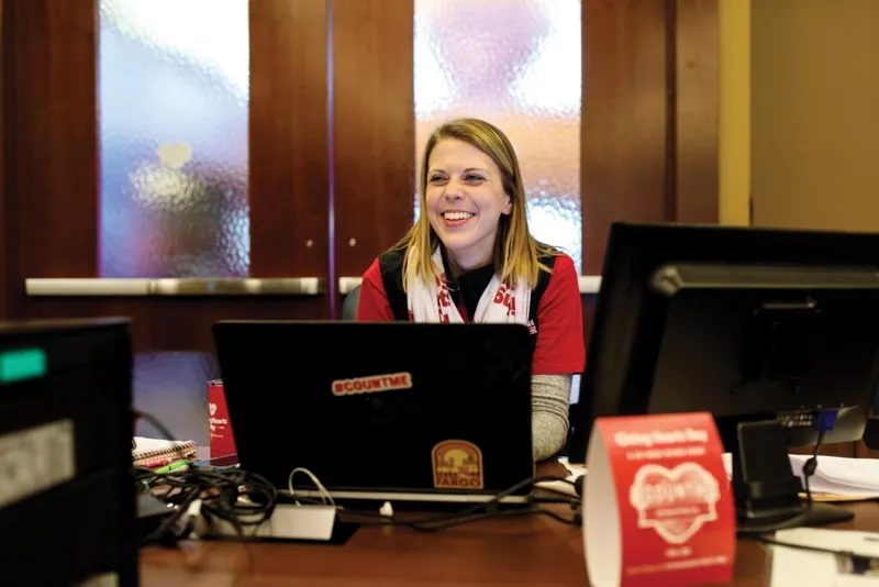 Smiling woman in red vest at a desk with a sticker-covered laptop during a giving event