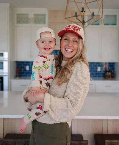 Smiling woman in a red trucker hat holding a laughing baby in printed pajamas in a bright white kitchen