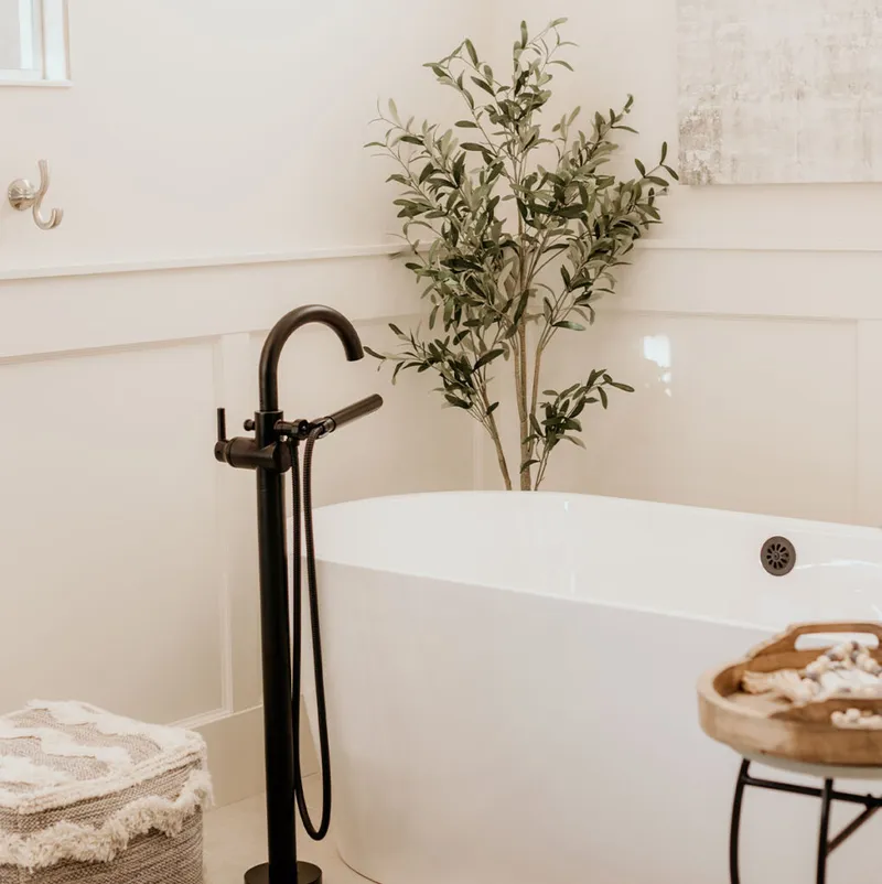 White freestanding soaking tub with a matte black floor-mount faucet and a tall leafy plant behind it