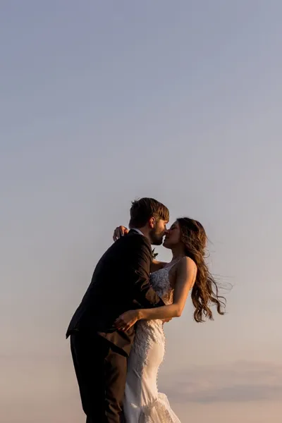 Bride in white lace gown and groom in dark suit kissing against a dusk sky