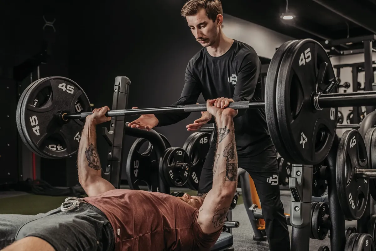 A spotter assists a tattooed man bench pressing a loaded 45-lb barbell in a dark gym