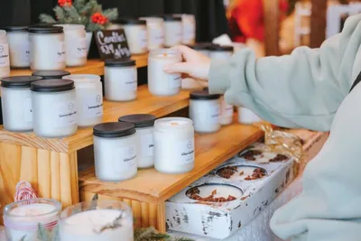 Person in light blue hoodie browsing rows of white candle jars on wooden market shelves