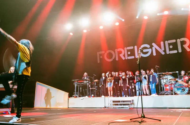 Foreigner concert with a singer onstage, arms raised, facing a large choir under red stage lights