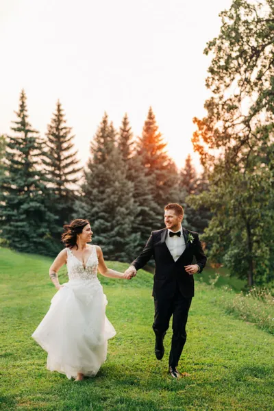 Bride in white gown and groom in black tuxedo holding hands running across a grassy lawn at sunset