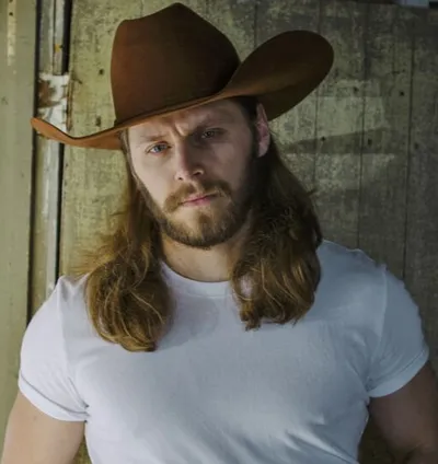 Bearded man with long auburn hair wearing a brown cowboy hat and white T-shirt