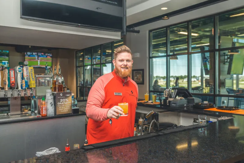 Smiling bearded bartender in red shirt holding an orange cocktail behind a bar
