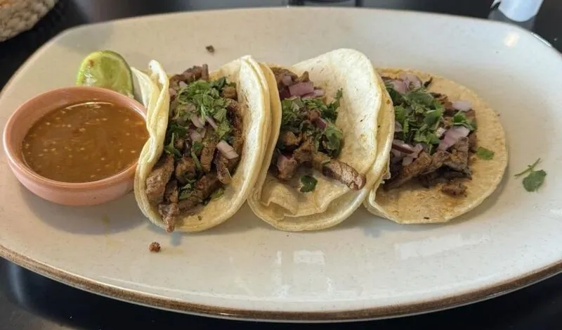 Three street tacos on corn tortillas topped with cilantro and red onion, served with salsa and a lime wedge