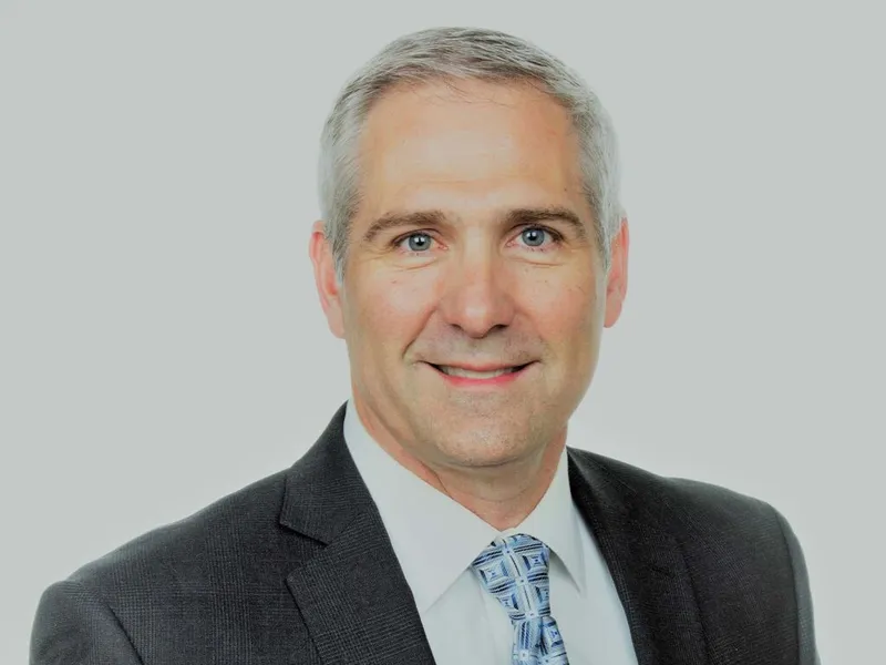 Man with gray hair in dark blazer and blue patterned tie smiling against light gray background