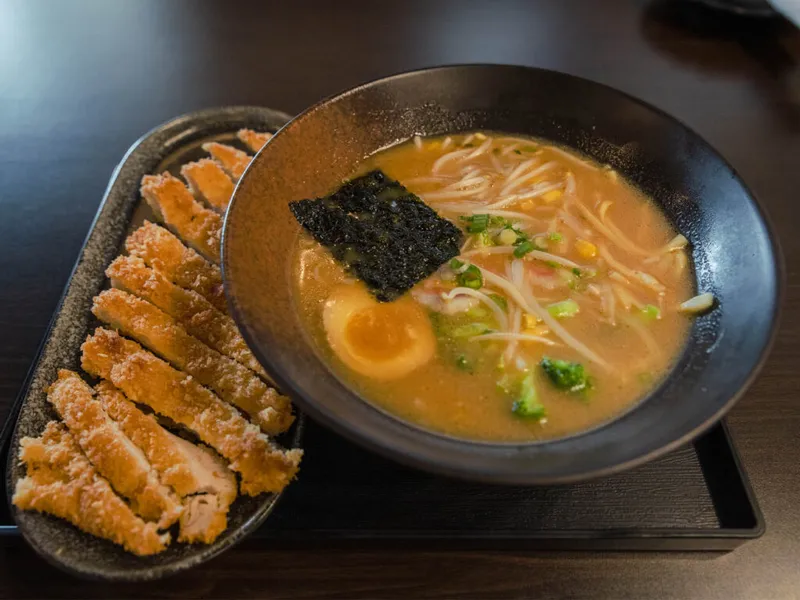 Bowl of miso ramen with egg, nori, broccoli, and bean sprouts beside sliced katsu on a dark tray