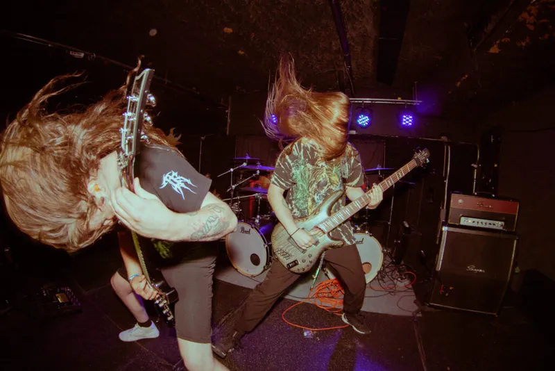 Two long-haired guitarists headbanging on a dimly lit club stage with blue stage lights