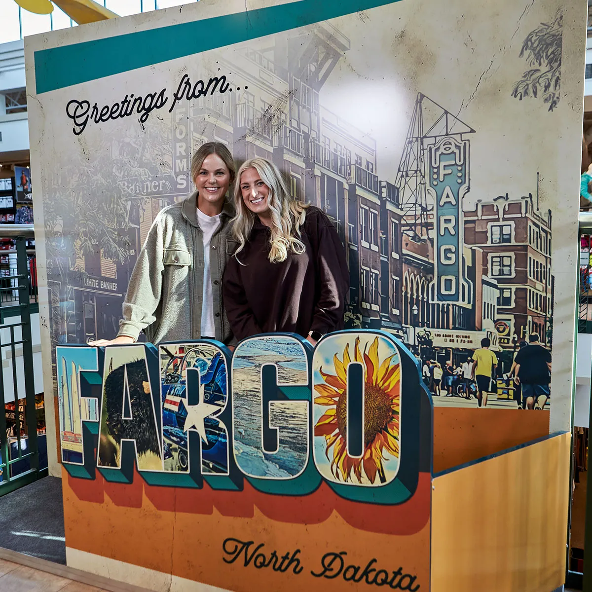 Two smiling women pose behind a large 'Greetings from Fargo, North Dakota' postcard display