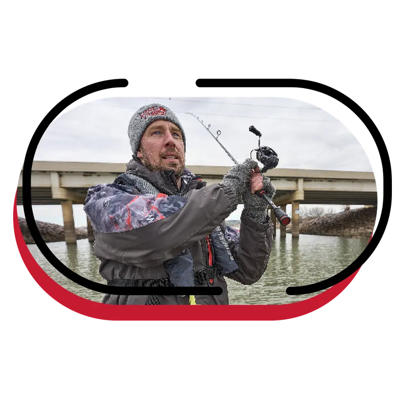 Man in winter gear and beanie holds a fishing rod near a bridge over calm water
