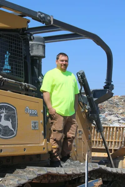 Man in neon yellow shirt standing on a large yellow bulldozer at a landfill