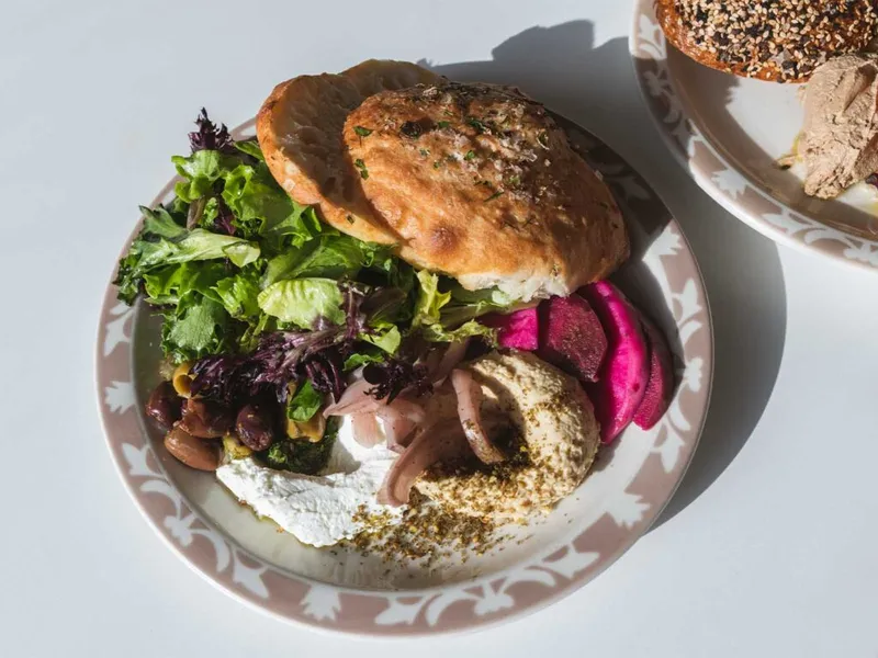 Patterned plate with a stuffed bread roll, mixed greens, hummus, olives, and pickled beets