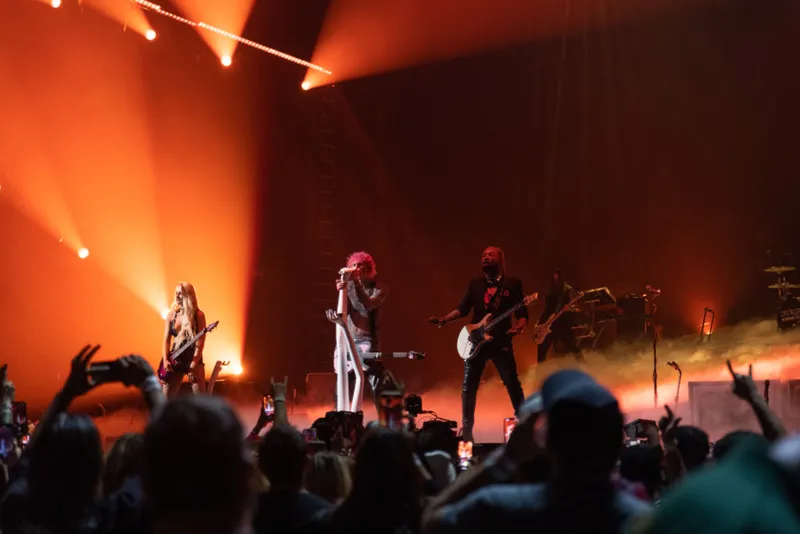Pink-haired performer on stage with two guitarists bathed in orange stage lighting before a packed crowd