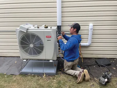 Technician in blue hoodie kneeling beside an outdoor HVAC unit mounted on a house exterior
